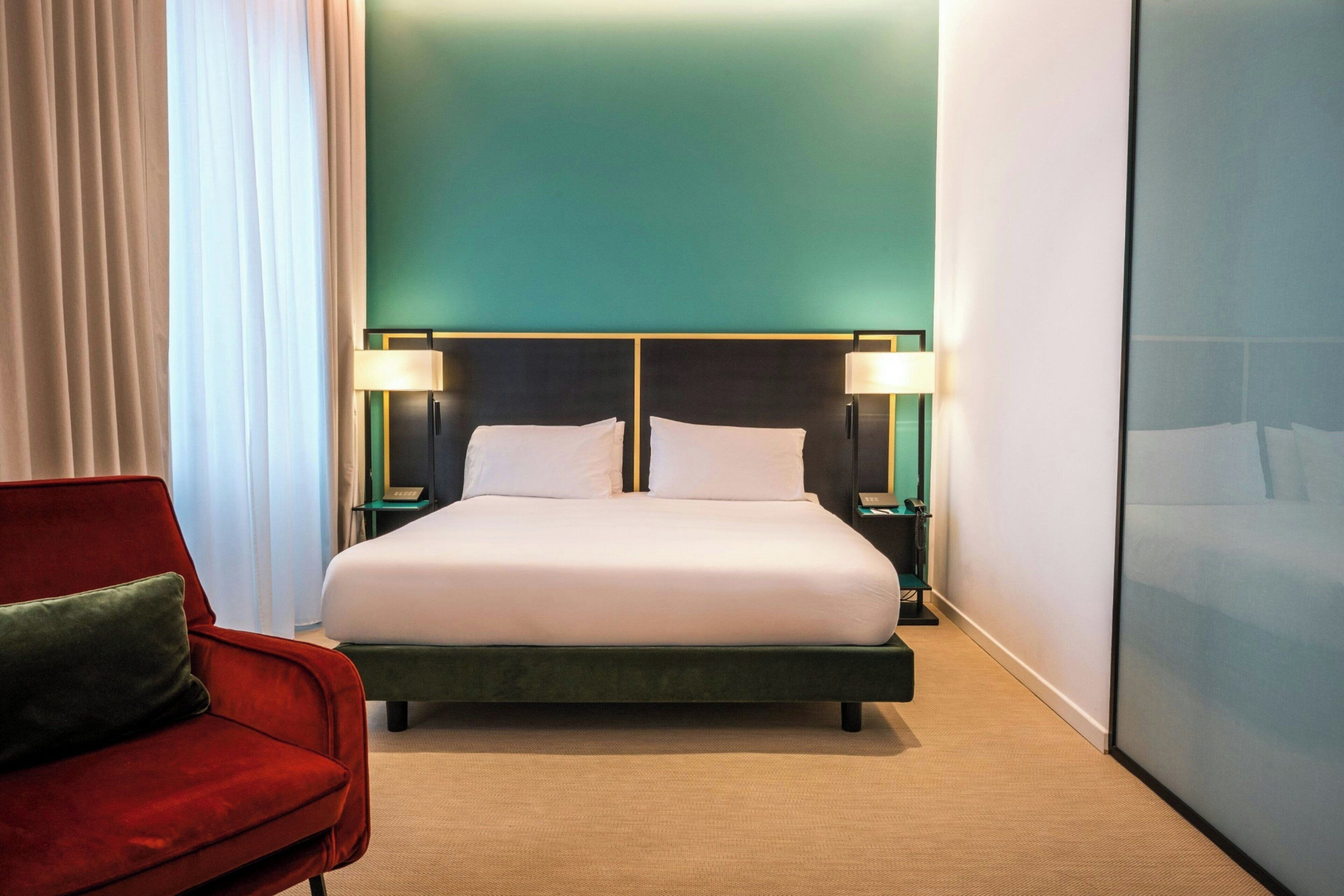 Een hotelkamer met een groot bed, twee nachtkastjes met lampen, een rode fauteuil en uitzicht op de stad.