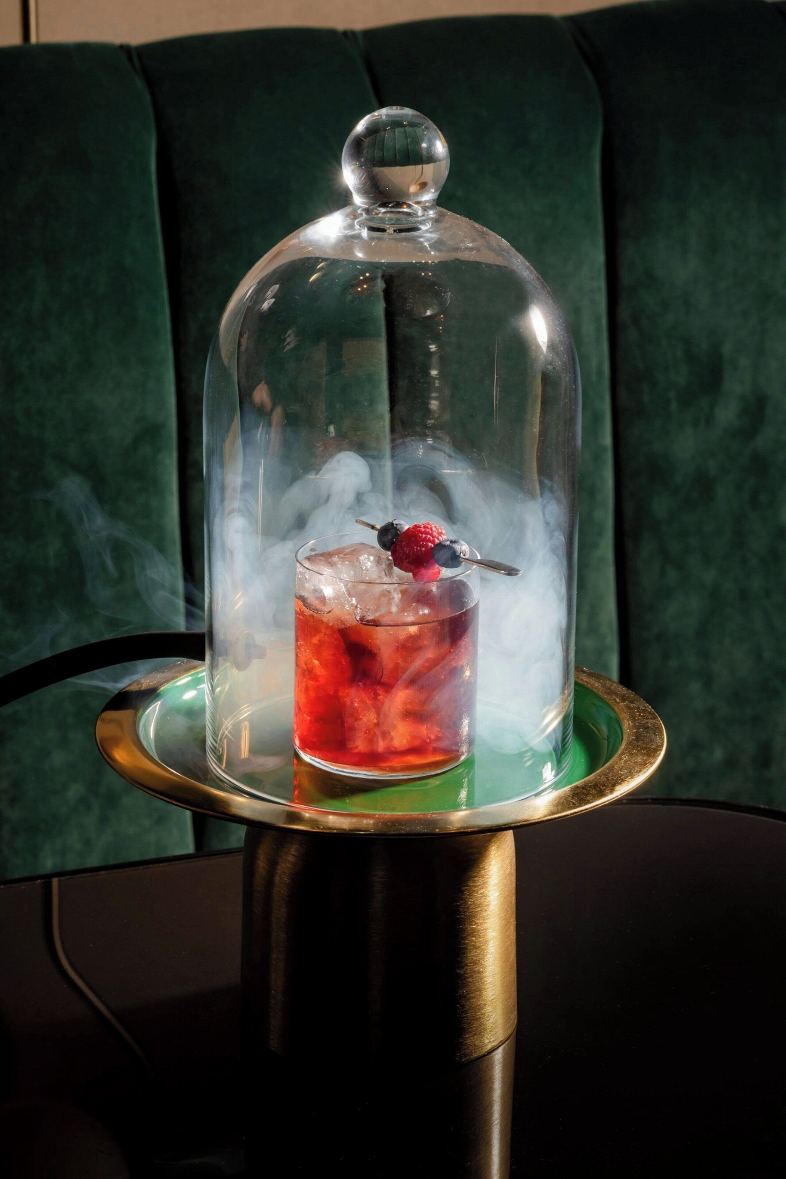 A glass of red beverage with ice and a berry garnish, under a glass dome, on a gold tray.