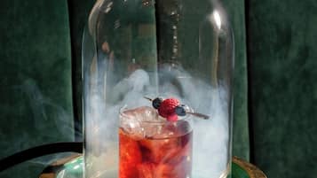 A glass of red beverage with ice and a berry garnish, under a glass dome, on a gold tray.