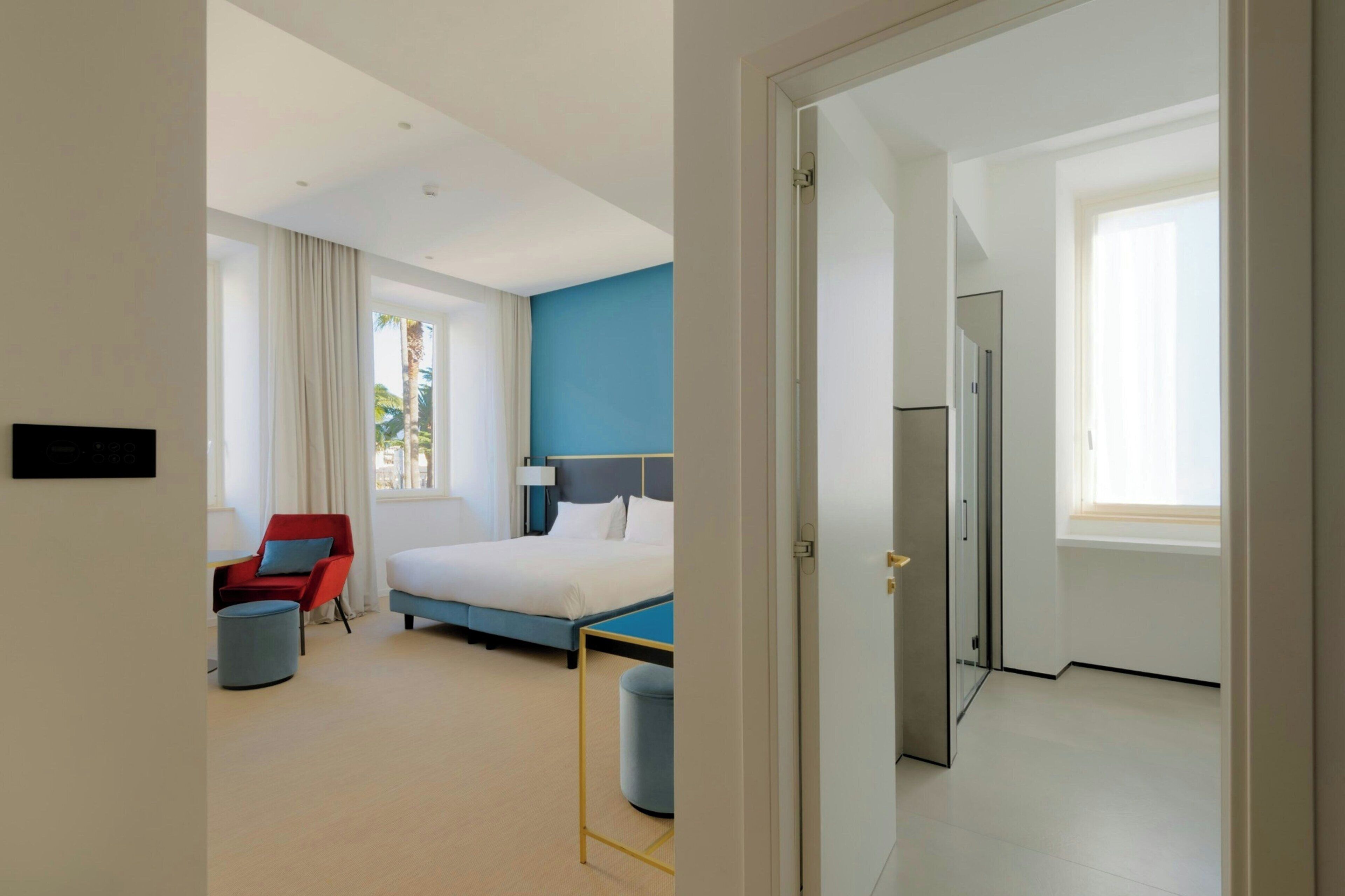 Una habitación de hotel con una cama, una silla roja, un reposapiés azul y una puerta que da a otra habitación.