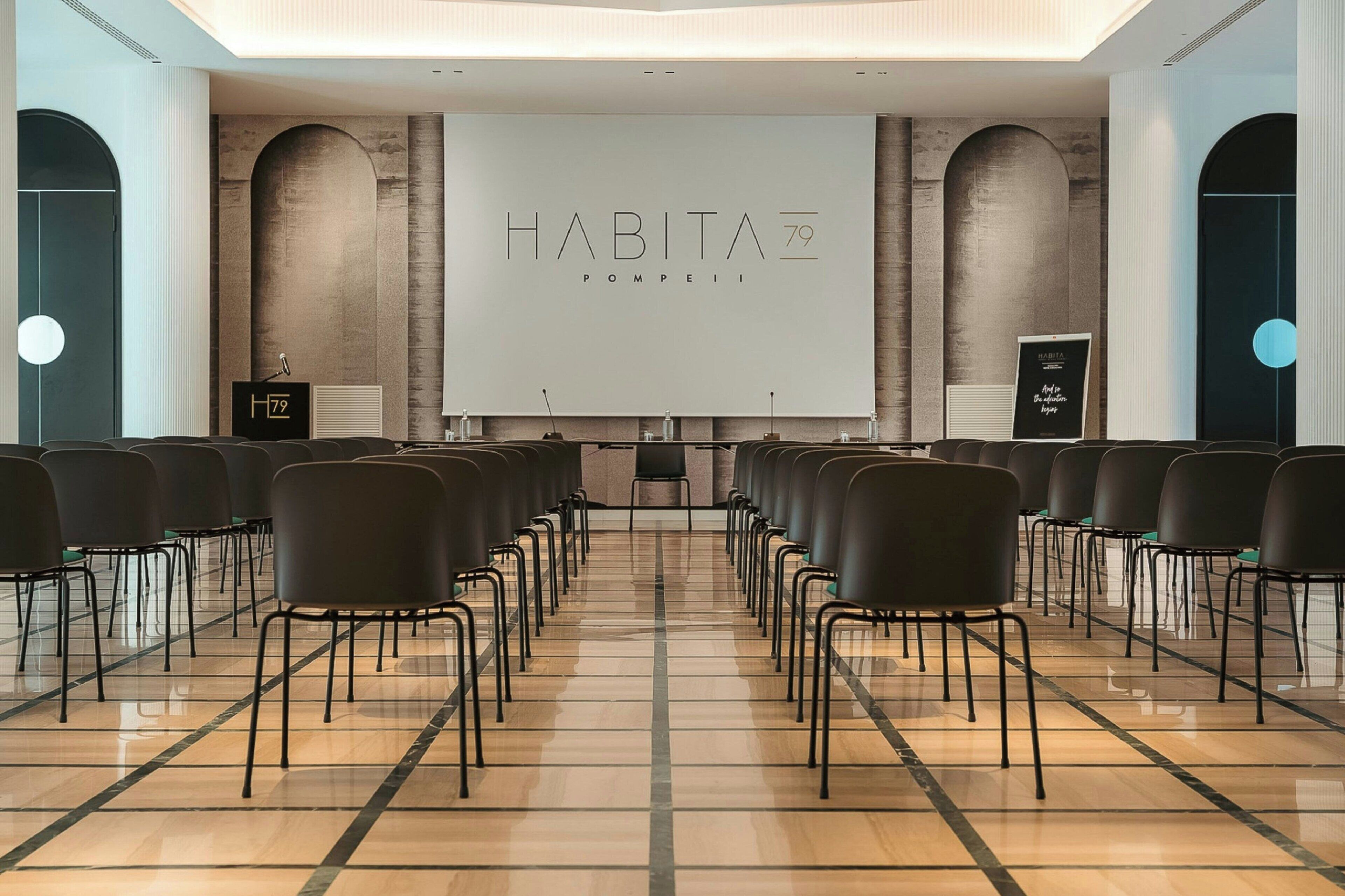 Un salón de conferencias con filas de sillas mirando hacia una pantalla grande que muestra 'HABITA 79 POMPEII'.