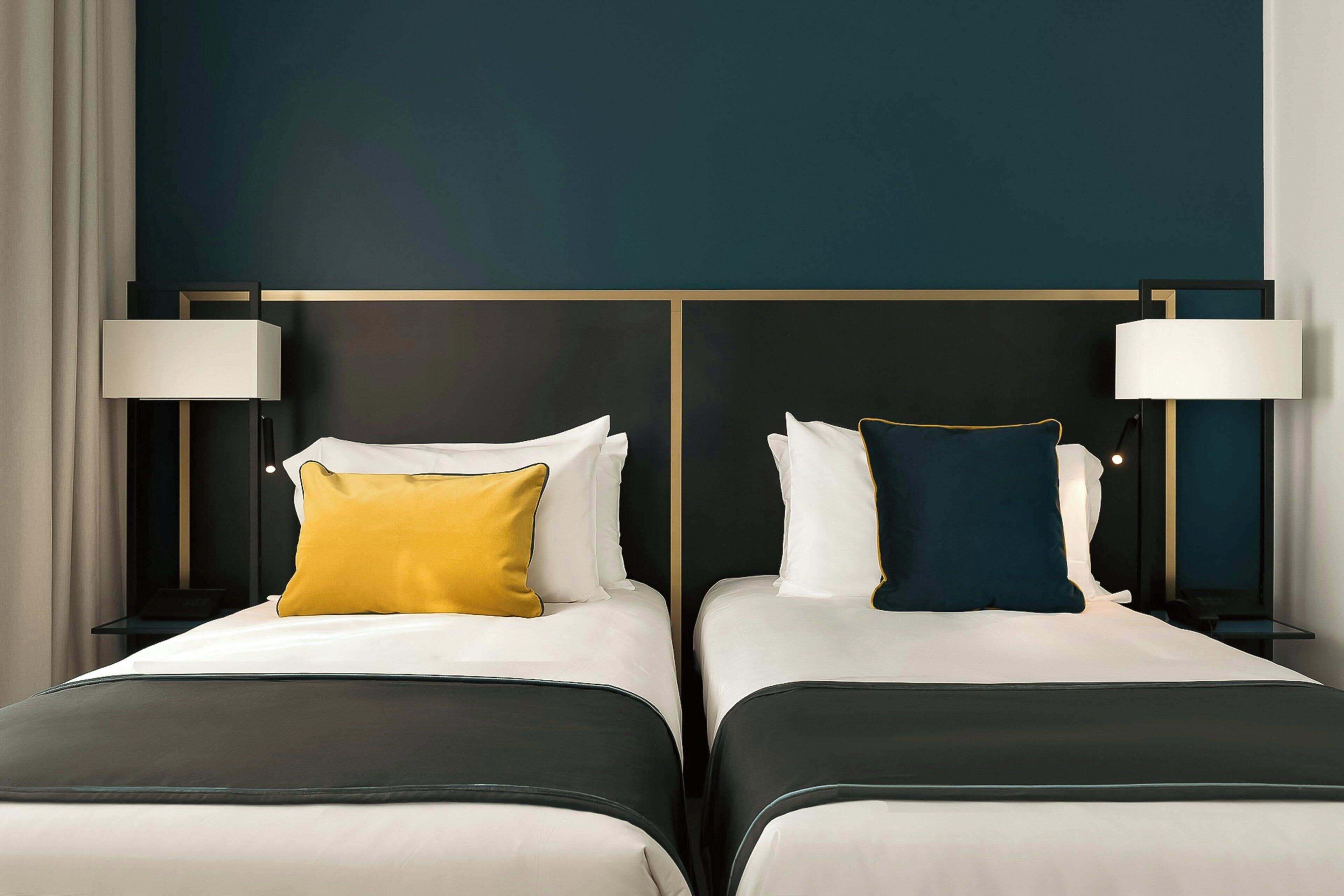 A hotel room with two beds, each with a nightstand, a lamp, and a pillow.