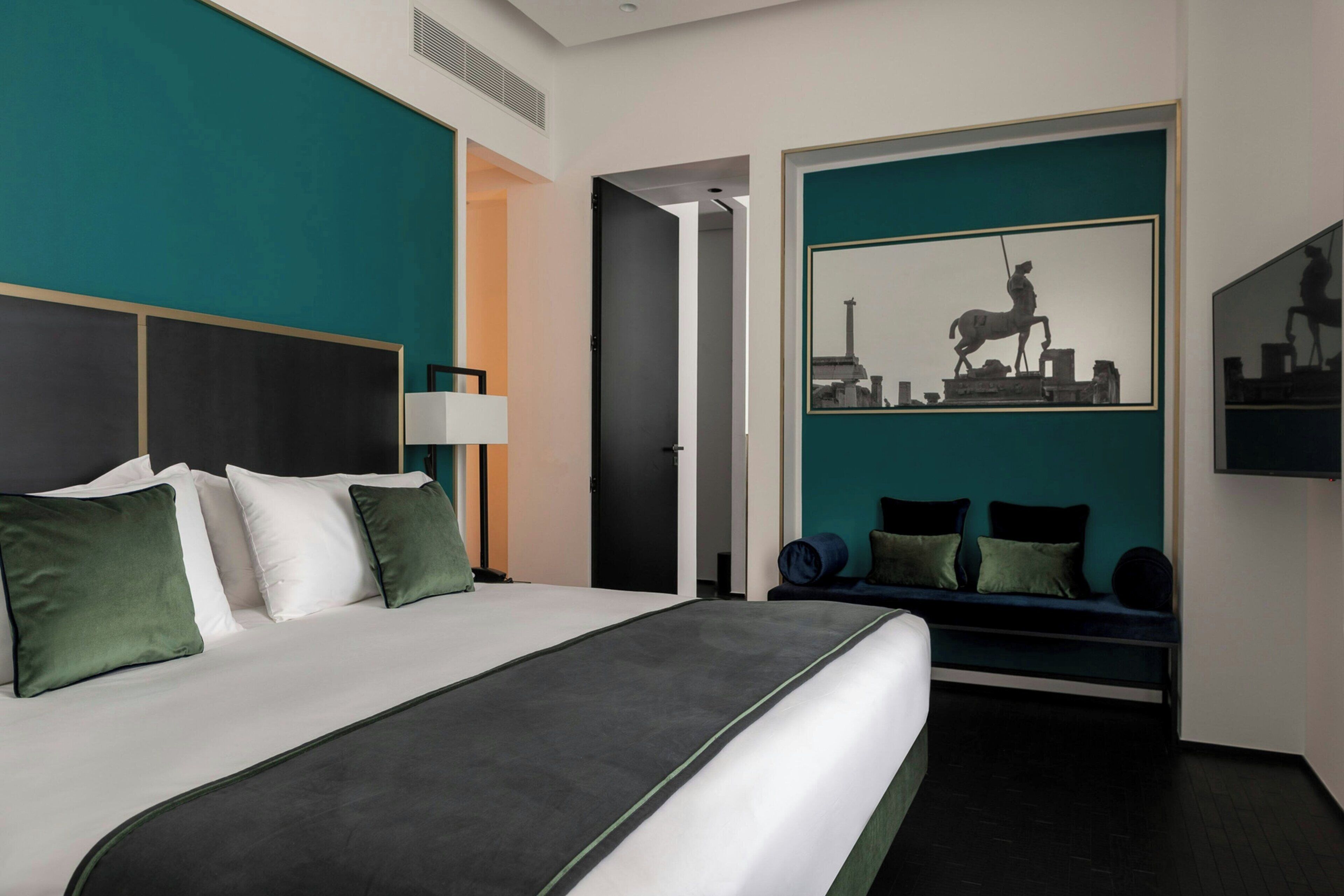 A modern hotel room with a large bed, a seating area with cushions, a painting of a horse statue, and a mirror on the wall.