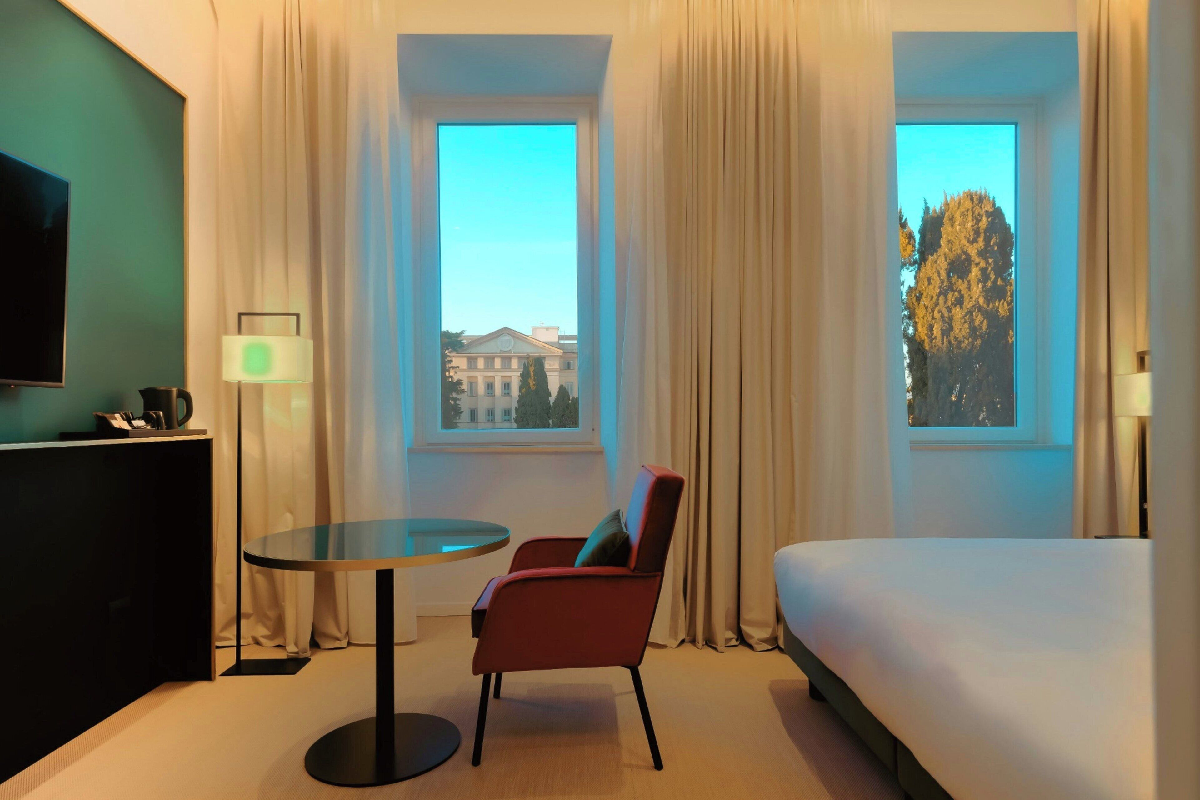 A hotel room with a bed, a red chair, a round table, a TV, and two windows with curtains.