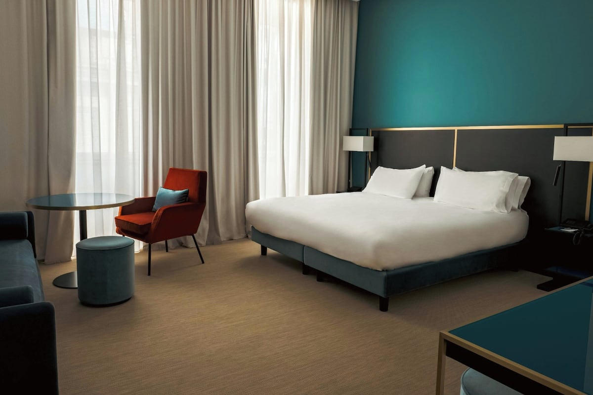 Quarto de hotel com uma cama, uma mesa redonda, uma cadeira vermelha, um banquinho azul e uma mesinha de cabeceira com abajur.