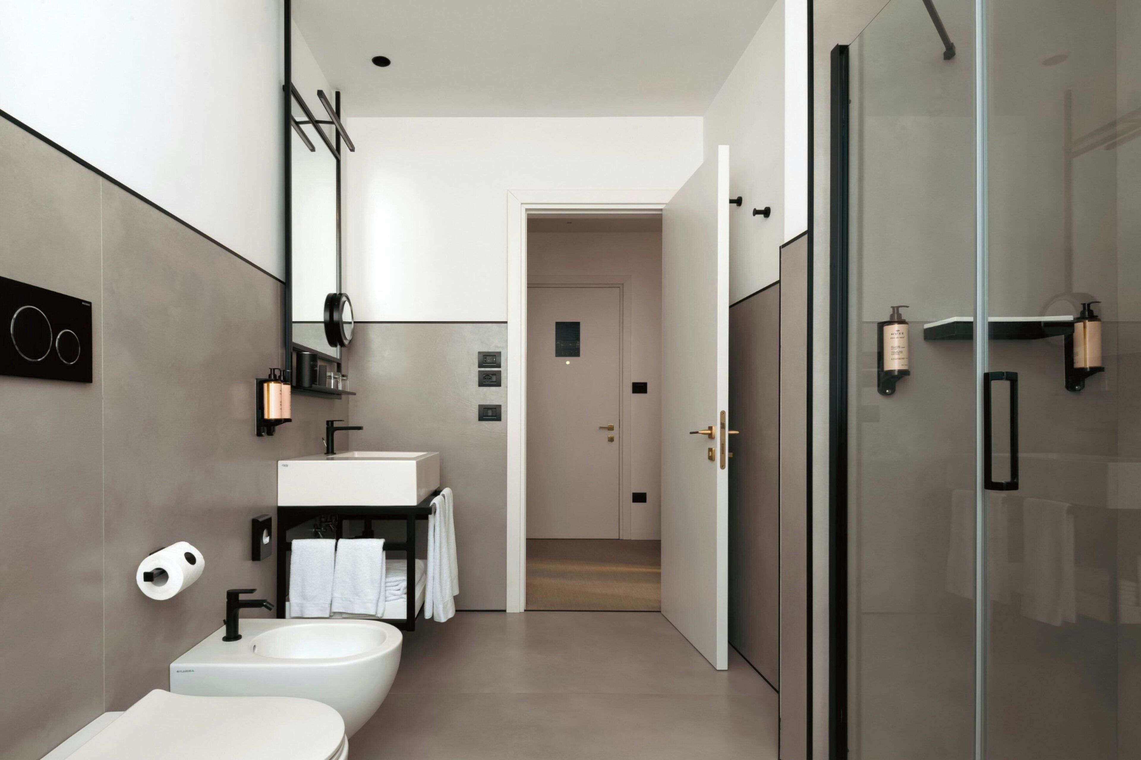 A modern bathroom with a toilet, bidet, and sink, a glass shower enclosure, and a hallway leading to another room.
