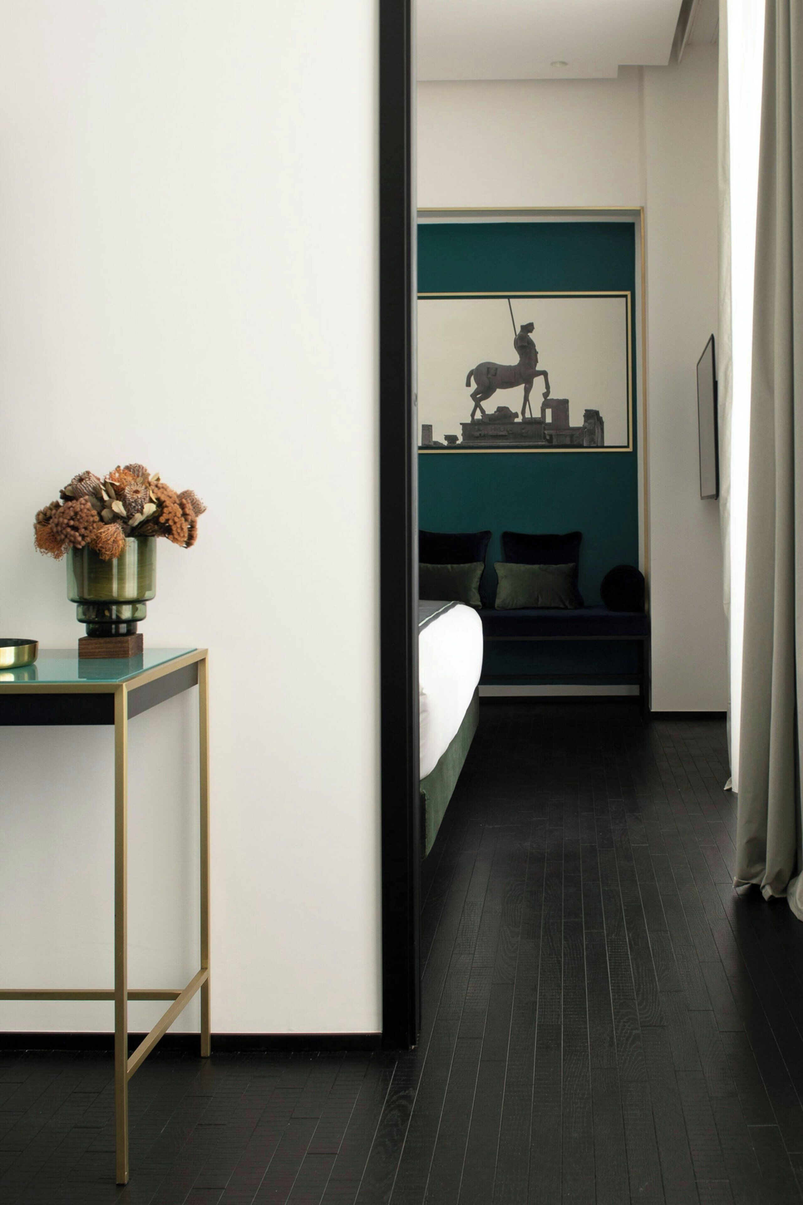 A modern hotel room with a dark wooden floor, a glass table with a vase, a bed with dark bedding, and a teal accent wall with a framed picture.