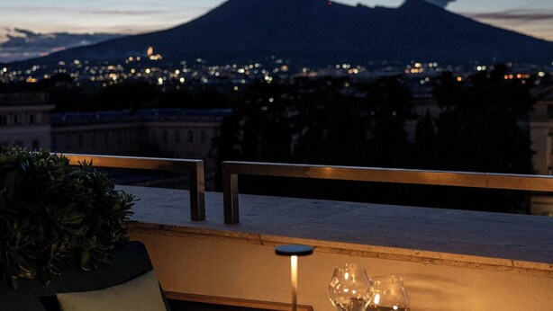 A table set for dining with glasses and a candle, overlooking a cityscape and a mountain at dusk. - HABITA79, Pompeii, a Tribute Portfolio Hotel (Pompei)
