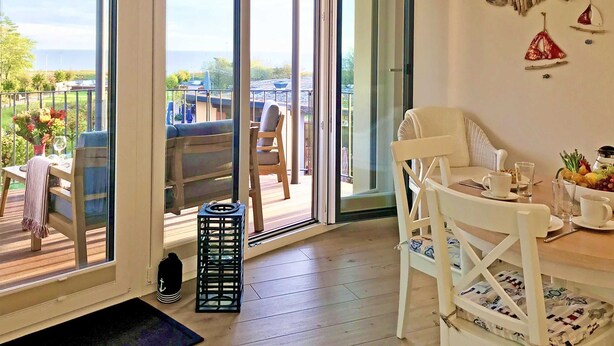 Una habitación con puerta corrediza de vidrio que da a un balcón con mobiliario exterior. Se observa una pequeña mesa con una silla, una silla blanca con cojín y un pequeño cúbito negro.