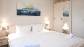 Una cama bien hecha con sábanas y almohadones blancos, una toalla doblada sobre la cama y un cuadro en la pared superior.