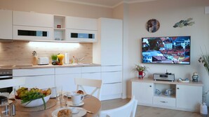 Una cocina moderna con armarios blancos, un comedor con una mesa preparada para dos personas y un televisor de pantalla plana montado en la pared que muestra un paisaje urbano.