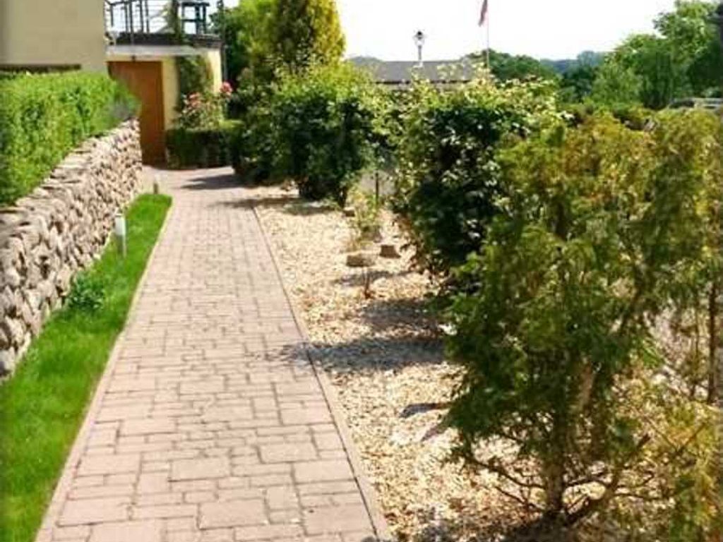 A paved pathway flanked by greenery and a stone wall, leading to a building.