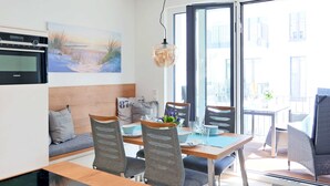 A modern kitchen with a built-in oven, a dining area with a wooden table and grey upholstered chairs, and a large window offering a view of the outside. - Baabe Villen Ensemble FeWo 15 Strandsonne Ref. 213134 (Baabe)