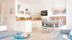 A modern kitchen with white cabinets, a dining area with a wooden table and chairs, and a view of the beach through a large window. - Baabe Villen Ensemble FeWo 15 Strandsonne Ref. 213134 (Baabe)