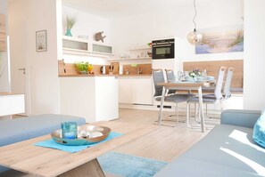 Uma cozinha moderna com armários brancos, um espaço de jantar com mesa e cadeiras de madeira, e vista para a praia através de uma grande janela.