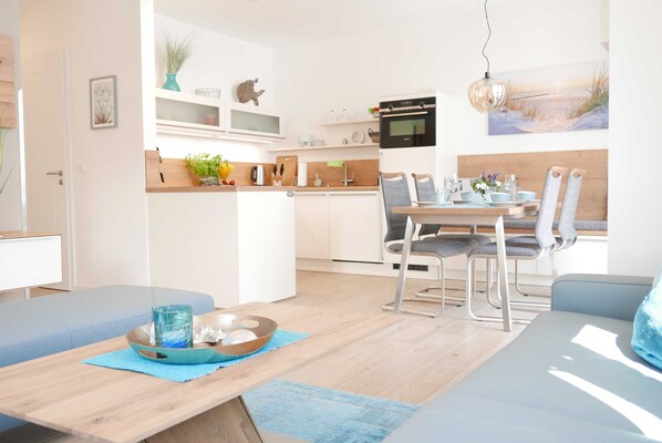 A modern kitchen with white cabinets, a dining area with a wooden table and chairs, and a view of the beach through a large window. - Baabe Villen Ensemble FeWo 15 Strandsonne Ref. 213134 (Baabe)