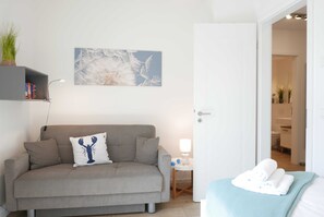 Quarto de hotel moderno com sofá cinzento, prateleira na parede com livros, pintura decorativa de dente-de-leão, mesa de cabeceira com abajur e cama bem arrumada com toalha dobrada.