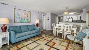 A living room with a blue sofa, a dining table with white chairs, and a kitchen area in the background. - Sand Pointe - Oceanfront Escape on 41st Street (Ocean City)