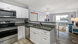 A modern kitchen with white cabinets, granite countertops, stainless steel appliances, and a large window overlooking a balcony. - Sand Pointe - Oceanfront Escape on 41st Street (Ocean City)
