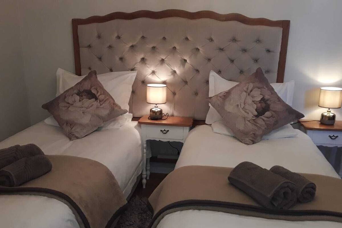 A hotel room with two beds, each with a pillow and a rolled towel. There is a nightstand with a lamp between the beds.