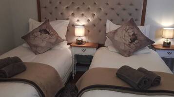 Een hotelkamer met twee bedden, elk met een kussen en een opgerolde handdoek. Tussen de bedden staat een nachtkastje met een lamp.