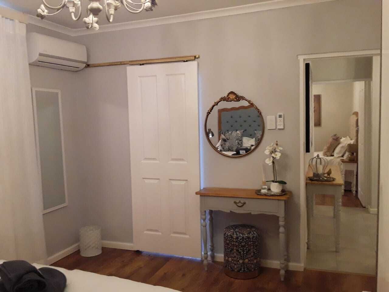 A bedroom with a wooden floor, a white door, a vanity with a mirror, and a small table with a potted plant.