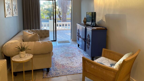 A living room with a sofa, a chair, a small table with a plant, a blue cabinet with decorative items, and a view through a glass door to an outdoor area. - Dellville Country House (Kareeberg)