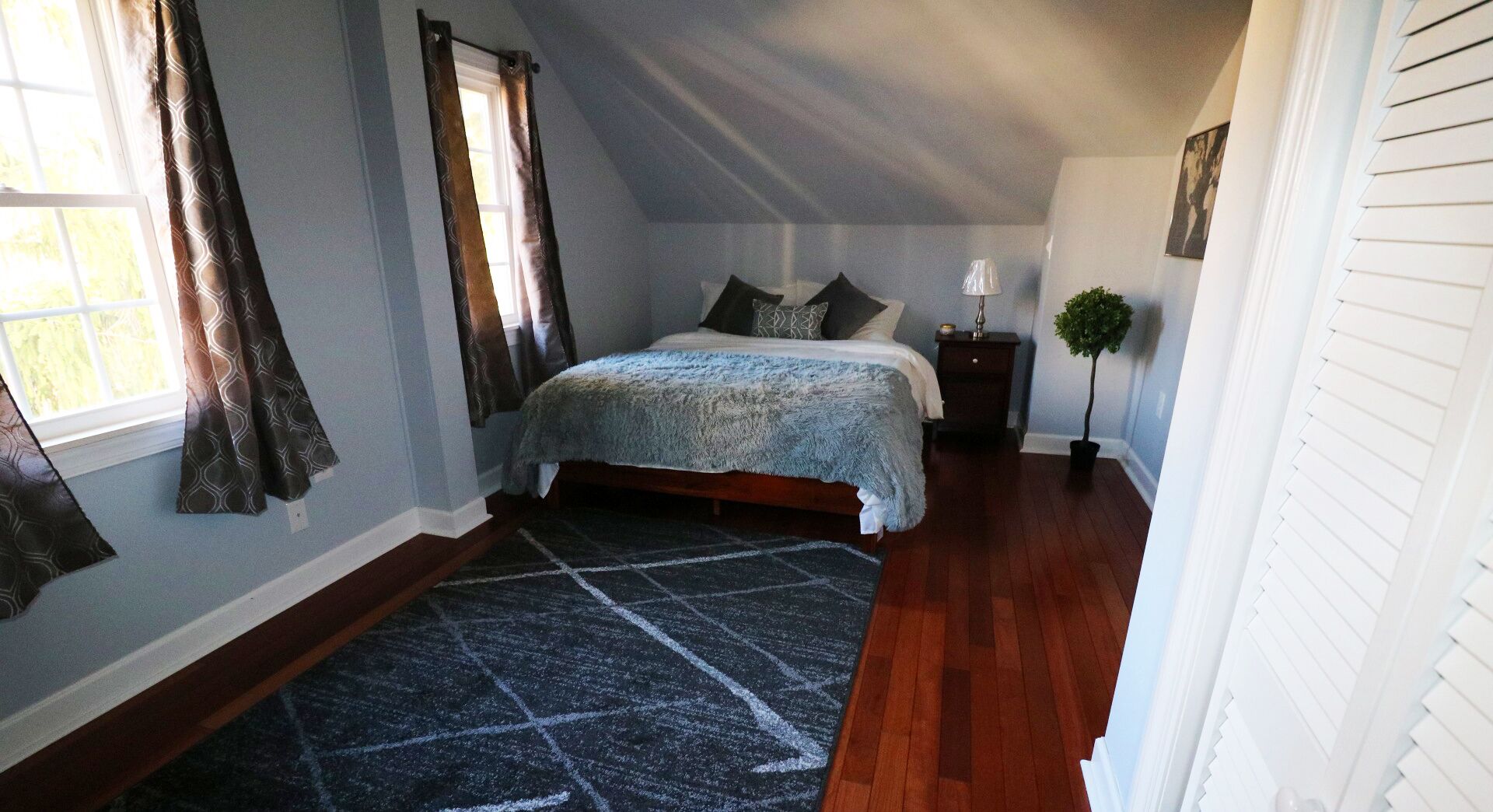 Un dormitorio con una cama, una mesita de noche, una lámpara, una planta en maceta y un ventanal con cortinas.