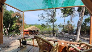 A wooden deck with wicker chairs and a table, overlooking a thatched-roof hut and a lake. - 4k Camp Koh Rong (Koh Rong)