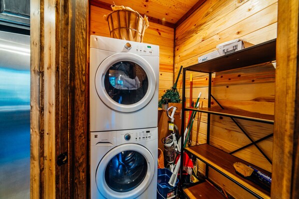 A compact laundry area with a front-loading washer and dryer, wooden walls, and a metal shelf with cleaning supplies.