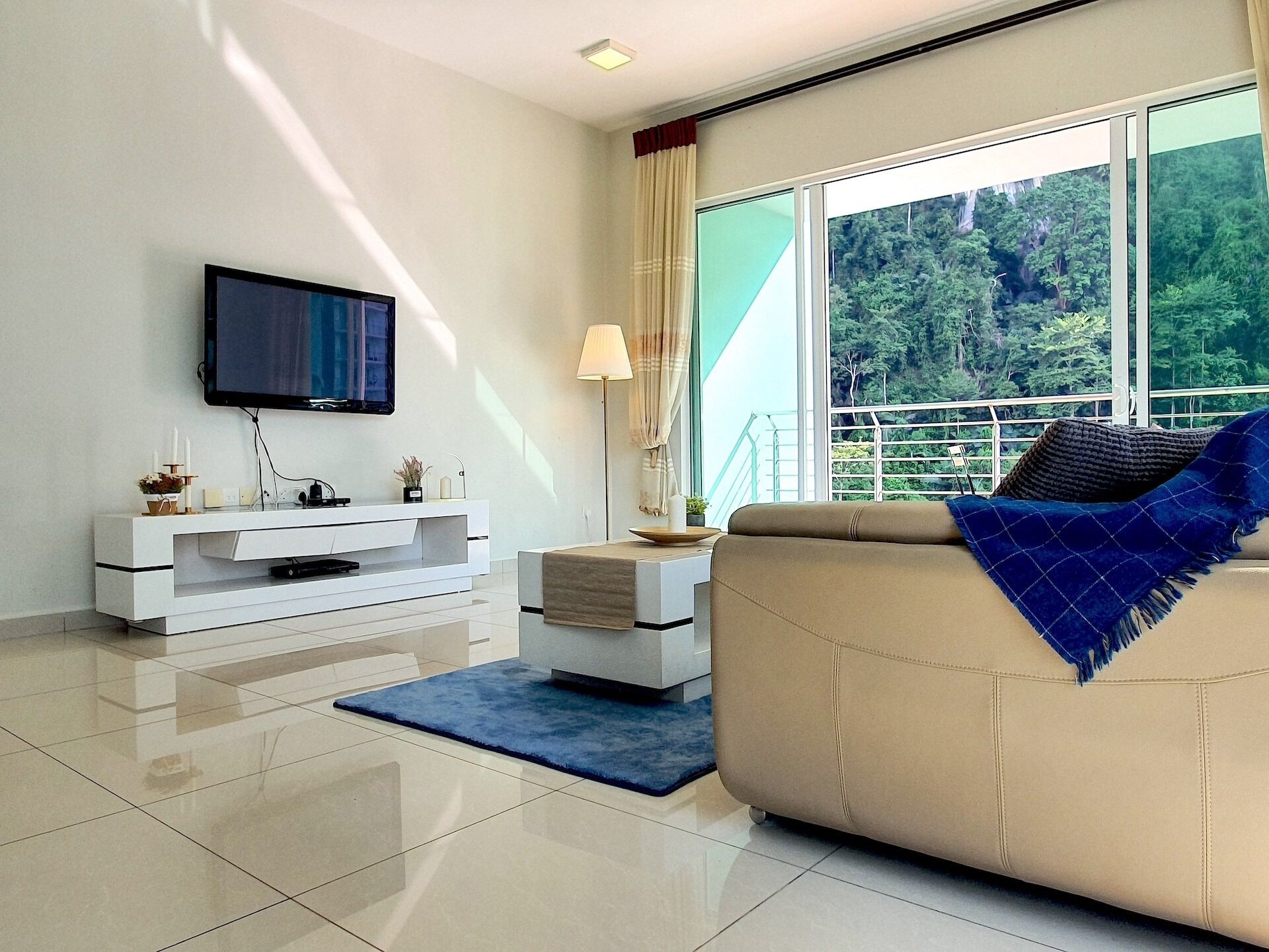 A modern living room with a flat-screen TV, a white entertainment unit, a beige sofa with a blue throw, and a large window with a view of greenery.