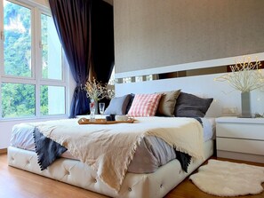 A modern bedroom with a large bed, a bedside table with a vase of flowers, and a view of greenery outside.