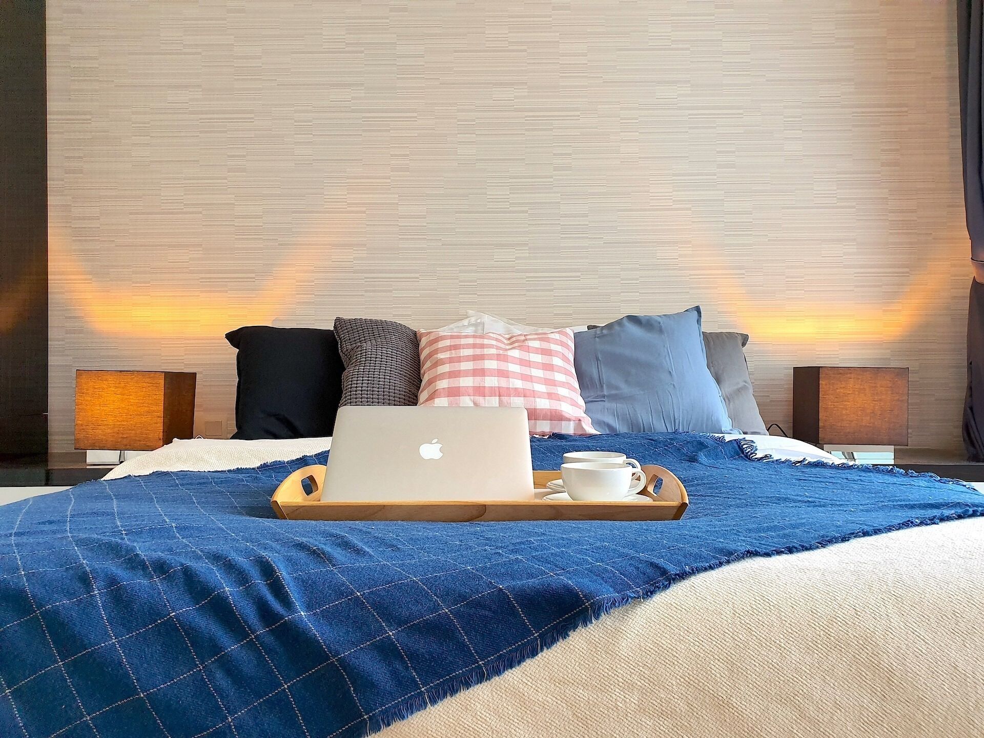 A neatly made bed with a laptop, a tray with cups, and a blue blanket.