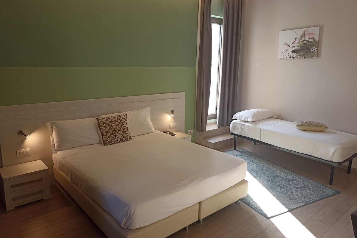 Ein Schlafzimmer mit einem großen Bett, einem kleineren Bett, einem Nachttisch, einer Fensterbank und einem Kunstwerk an der Wand.