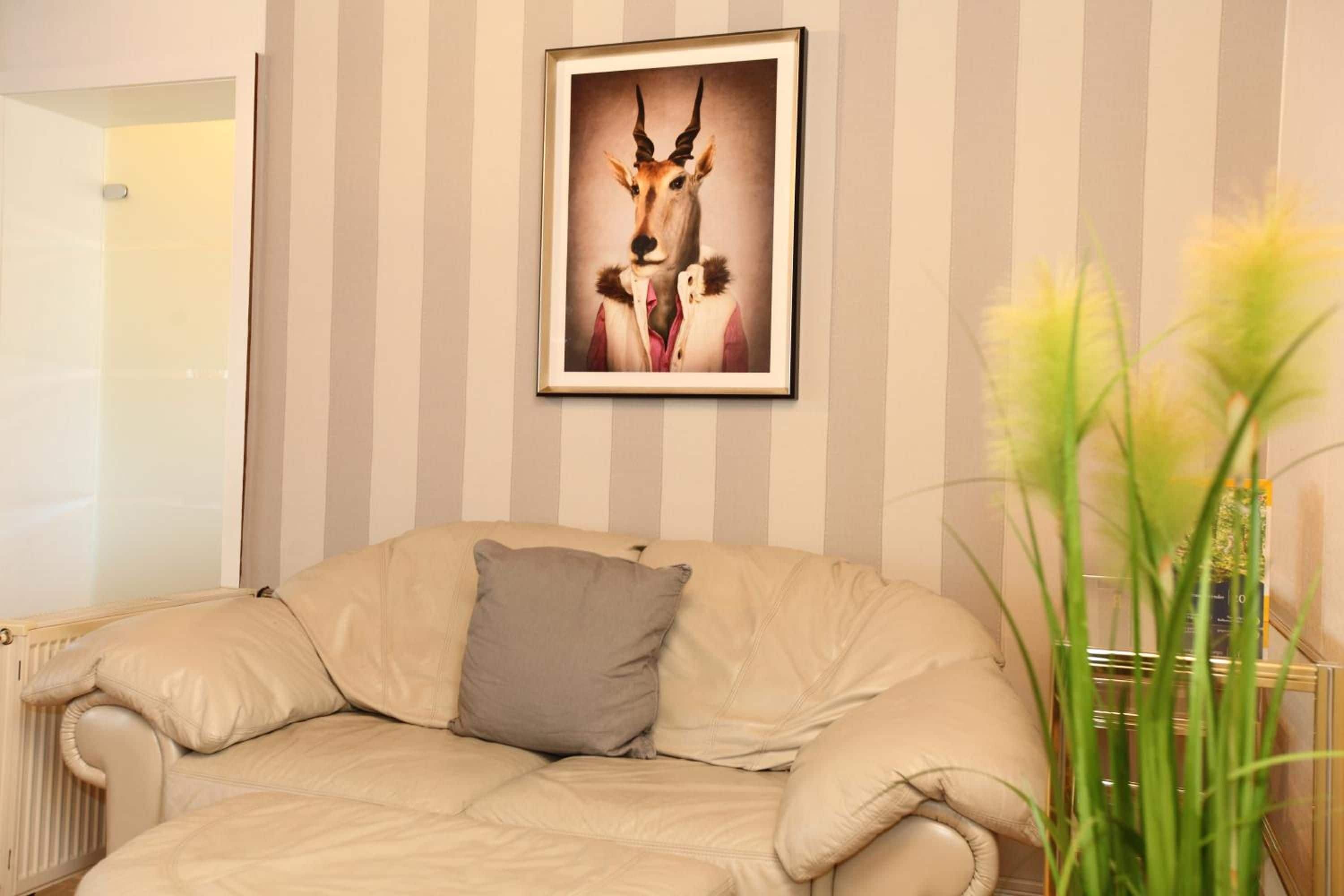 A room with a striped wall, a framed picture of an animal, a beige sofa with a pillow, and a plant.