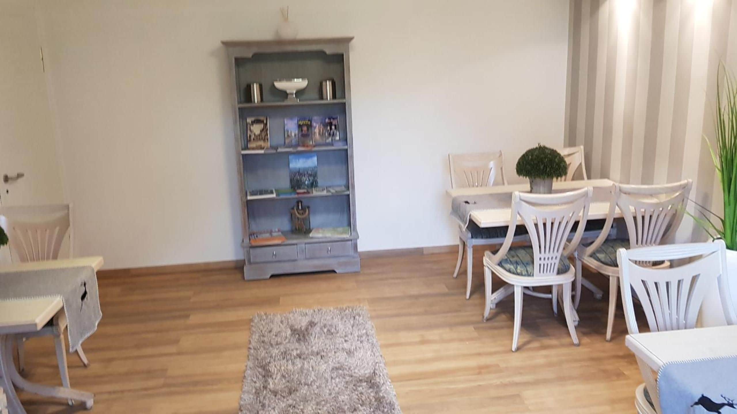 Una sala de estar con piso de madera, una estantería gris, sillas blancas, una mesita y un tapete gris.