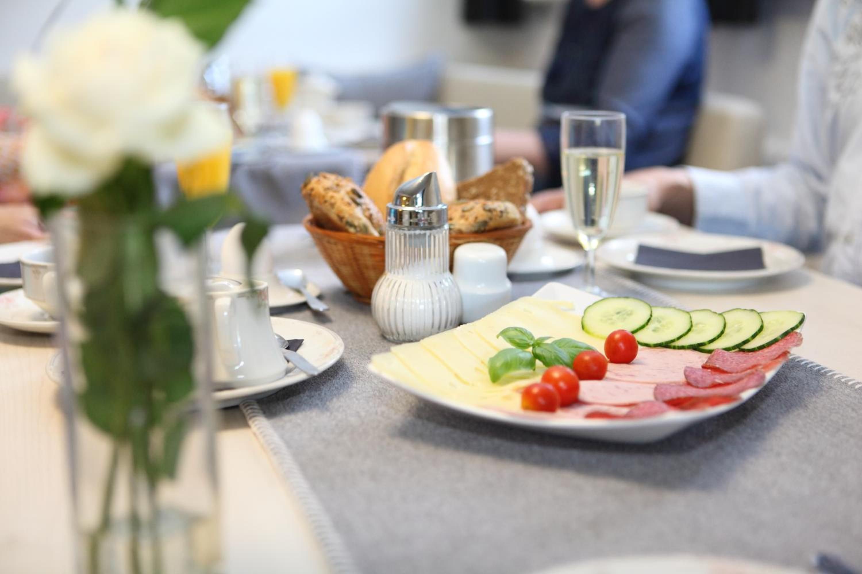 Een ontbijttafel met een bord gesneden komkommers, tomaten en kaas, een broodmand, een peper- en zoutstel en een glas water.