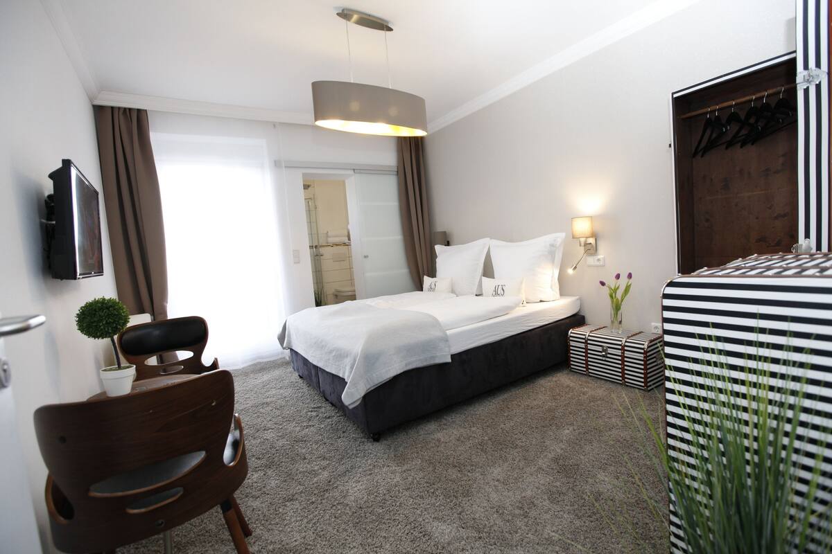Een moderne hotelkamer met een groot bed, een bureau met stoel, een kleine plant en een kledingkast.