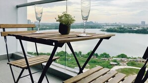 Une table en bois, avec deux verres et une plante en pot, sur un balcon donnant sur une rivière et la ville.