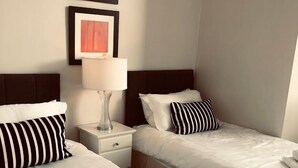 Une chambre d’hôtel avec deux lits, chacun étant équipé d’un oreiller et d’une couverture. Une table de chevet avec une lampe se trouve entre les deux lits.