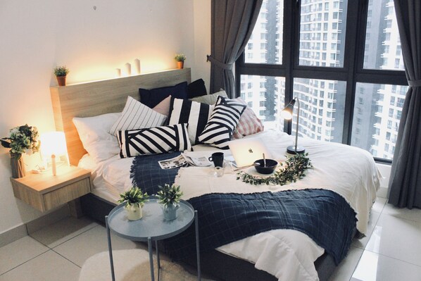 Un dormitorio ordenado con una cama grande, mesitas de noche, una pequeña mesa redonda y vistas de edificios altos a través del ventanal.