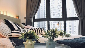 Ein Schlafzimmer mit einem Bett, zwei Vasen mit Pflanzen, einem runden Tisch und Blick auf hohe Gebäude durch ein Fenster.