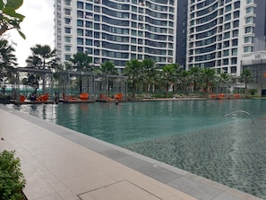 Una piscina con sillones de descanso y un área de estar cubierta, rodeada de palmeras y edificios modernos al fondo.