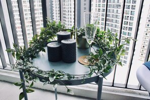 A round table with candles and a glass, surrounded by greenery, on a balcony with a city view.