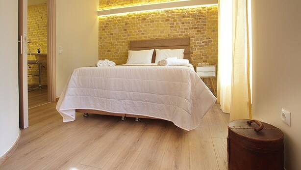 A neatly made bed with a headboard, a wooden floor, and a brick wall with a light fixture. - Liston Suite Plaza (Corfu)