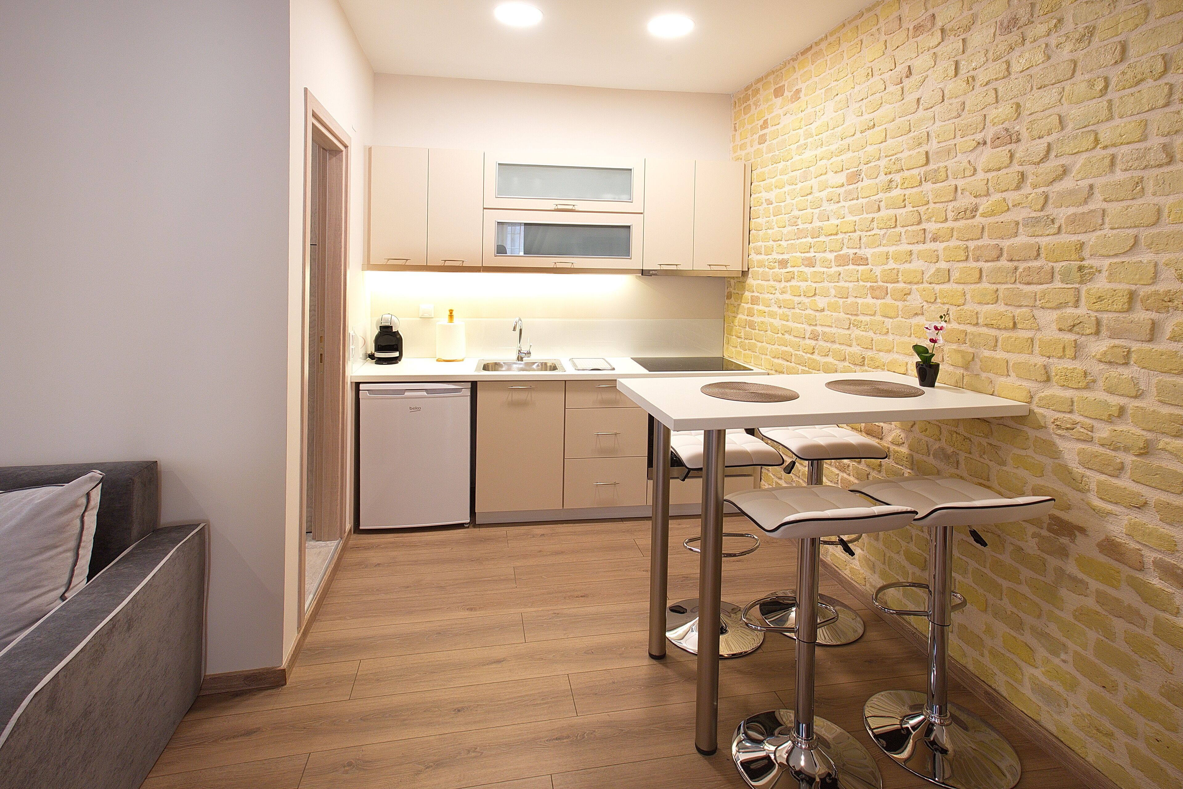 A compact kitchen with a wooden floor, a dining table with stools, and a brick wall.