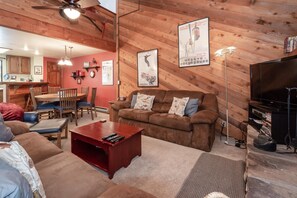 A cozy living room with a brown sofa, a wooden dining table, and a red coffee table. There is a television on the right and a ceiling fan above.
