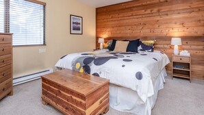 A bedroom with a wooden headboard, a large bed with a white and black patterned duvet, two bedside tables with lamps, a wooden chest of drawers, and a window with blinds.