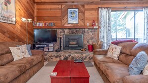 A cozy cabin interior with two brown leather sofas, a stone fireplace, a television, and a red coffee table.