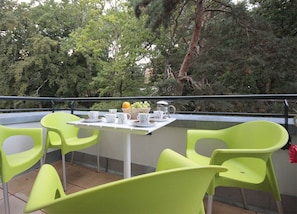 Un balcon avec une table dressée pour deux personnes, entouré de chaises vertes, et un panier de fruits.